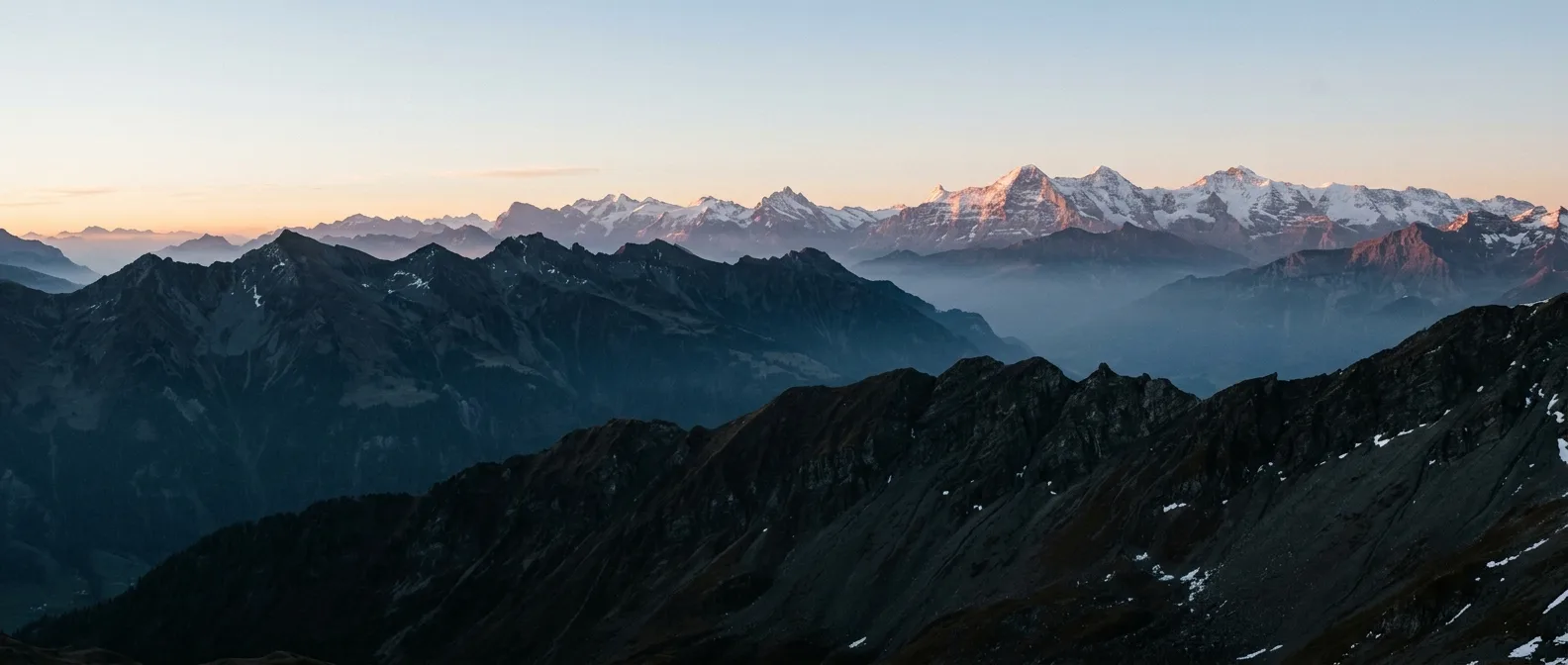 Layered Bernese Oberland ridgelines receding into cold haze at first light, the highest snowfields catching warm alpenglow.