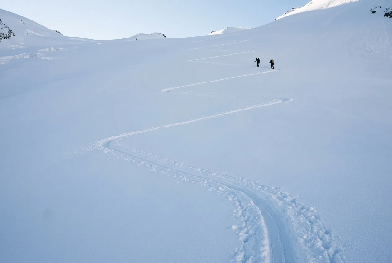 A skin track cutting long zigzags up an untracked snowfield, two small figures far up the slope.